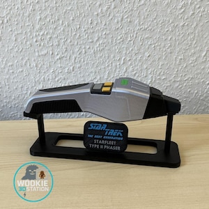May include: A silver and black Star Trek Type II Phaser replica, with yellow and green accents, displayed on a black stand. The stand also holds a black plaque with the Star Trek logo and text.