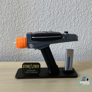 Könnte beinhalten: Eine Star Trek Starfleet Typ II Phaser-Nachbildung in Grau und Schwarz mit orangefarbener Emitterspitze, montiert auf einem schwarzen Ständer. Der Ständer hat ein Schild mit der Aufschrift STAR TREK STARFLEET TYPE II PHASER und ein silbernes Bauteil.