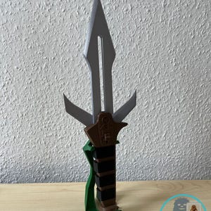 Könnte beinhalten: Ein 3D-gedruckter Dolch mit silberner Klinge und braun-schwarzem Griff. Der Griff hat einen grünen Sockel und ein grünes Band. Der Dolch wird vor einer strukturierten weißen Wand präsentiert.