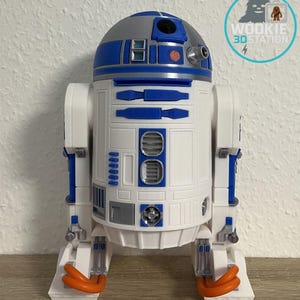 Könnte beinhalten: Ein detailliertes 3D-gedrucktes Modell von R2-D2, dem ikonischen Astromech-Droiden. Das Modell hat einen weißen Körper mit blauen und silbernen Akzenten, einschließlich der charakteristischen blauen Paneele und der Kuppel. Orangefarbene Fußdetails runden das Design ab.