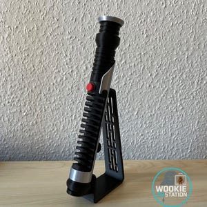 Puede incluir: Un sable de luz negro con detalles plateados y un botón rojo, exhibido en un soporte negro. El sable de luz tiene un mango acanalado y una base plateada. El soporte está inclinado y tiene cortes verticales.