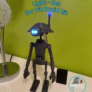 Op de afbeelding: Een donkergrijze Pit Droid-kit met een lichtset, met een blauw oplichtend oog en antenne. De tekst "Light - Set for Pit Droid kit" staat boven de droid. Een zwart batterijpakket staat naast de droid.