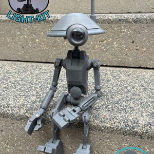 May include: A gray articulated robot figure with a domed head, large eye, and antenna. The figure is sitting on a concrete surface. The image includes the text "compatible LIGHT-KIT" and the "Wookie Station" logo.