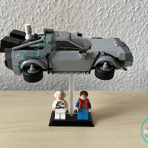 May include: A gray Lego DeLorean time machine model with black wheels and a clear stand. Two Lego figures, one with white hair and the other with brown hair, stand on a black base. The image includes a logo that says "Wookie 3D Station".