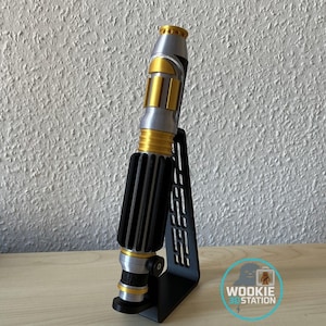Peut inclure: Un sabre laser argenté, doré et noir, présenté sur un support noir. Le sabre laser présente une poignée noire côtelée, des détails dorés et un émetteur argenté. Le support a un design découpé. Le logo Wookie 3D Station est visible.