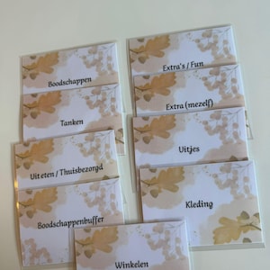Op de afbeelding: Een verzameling gelabelde kaarten in doorzichtige plastic hoezen. Elke kaart heeft een aquarelontwerp van eikenbladeren en bessen in bruin- en goudtinten. Tekst op de kaarten is onder meer "Extra's/Fun", "Boodschappen" en "Winkelen".