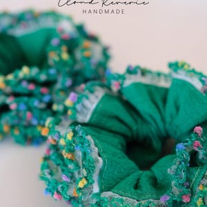 May include: Two green scrunchies with colorful, textured trim. The scrunchies are made of a textured fabric and have a white lace trim. The text "Cloud Reverie HANDMADE" is visible in the background. These hair accessories are perfect for adding a pop of color.