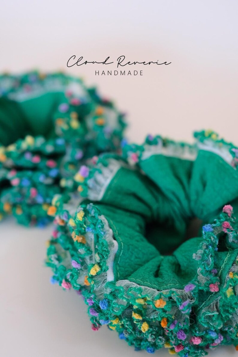 May include: Two green scrunchies with colorful, textured trim. The scrunchies are made of a textured fabric and have a white lace trim. The text "Cloud Reverie HANDMADE" is visible in the background. These hair accessories are perfect for adding a pop of color.