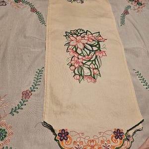 Può includere: Un runner da tavola rettangolare color crema con ricamo a macchina. Il centro presenta un gruppo di fiori rosa con steli e foglie verdi. Il bordo inferiore ha una decorazione smerlata con motivi floreali viola e bianchi.