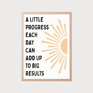 May include: A framed print with the text "A LITTLE PROGRESS EACH DAY CAN ADD UP TO BIG RESULTS" in black font. A peach-colored sun graphic is on the right side. The frame is a light brown color.