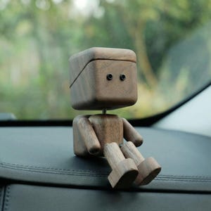 May include: A wooden figurine of a seated robot with a box-shaped head and small black eyes. The arms and legs are also made of wood. The robot is sitting on a dark surface, likely a car dashboard. The background is blurred, suggesting an outdoor setting.