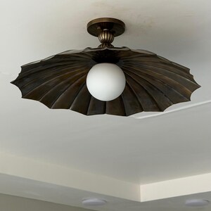 May include: A flush mount ceiling light fixture featuring a wide, dark bronze-toned metal shade with a scalloped, fan-like design. A central, spherical white glass globe bulb is suspended from a ribbed, cylindrical mounting base in matching bronze.