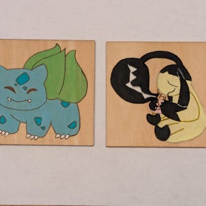 May include: Two square wooden tiles featuring hand-painted illustrations. The left tile displays a blue creature with a green bulb on its back. The right tile shows a yellow creature with black accents and a large black claw-like appendage.