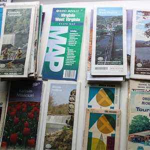 May include: A collection of vintage paper road maps from various states in the United States. The maps are arranged in a grid pattern on a white surface. The maps are all in different colors and have different designs. Some of the maps have the words "State Map" or "Tour Map" printed on them. The maps are all folded and have a worn look.