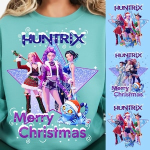 May include: Teal sweatshirt with a graphic featuring three stylized figures in festive outfits, a blue tiger wearing a Santa hat, and the text "Merry Christmas" and "Huntrix". Additional smaller graphics are on the right.