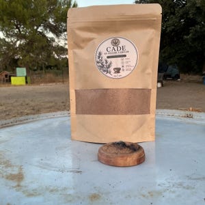 Peut inclure: Un sachet en papier kraft brun contenant de la poudre de cade à brûler, avec une étiquette circulaire portant le texte CADE EN POUDRE A BRULER. Un petit récipient en bois contenant un tas de poudre brune est posé devant le sachet.