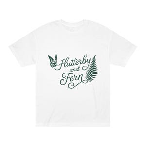 May include: White t-shirt with the text "Flutterby and Fern" in dark green script. A green butterfly and fern leaf are on the left and right of the text. The shirt is a casual, unisex style.