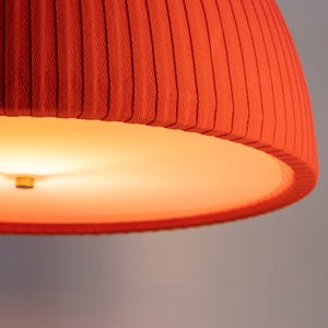 May include: Close-up of a red fabric lampshade with vertical pleats. The lampshade has a warm, orange glow emanating from the interior, creating a soft, inviting light. The design is modern and minimalist.