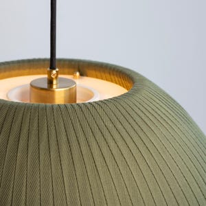 May include: Close-up of a green fabric pendant light fixture. The shade has vertical ribbed detailing. The fixture has a brass-colored metal base and a black cord.