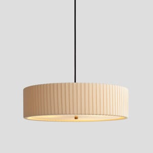 May include: A cylindrical pendant light fixture featuring a cream-colored, vertically pleated fabric shade. The drum-shaped shade is suspended by a single thin black cord and emits a soft, warm glow from the bottom opening of the structure.