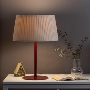 May include: A red table lamp with a pleated, cream-coloured lampshade. The lamp is on a white table, next to a small ceramic house and an open book. A glass vase with greenery is also on the table.