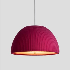 May include: A pendant light fixture with a deep magenta, pleated fabric shade. The shade is dome-shaped and hangs from a black cord. The interior of the shade has a warm, golden glow.