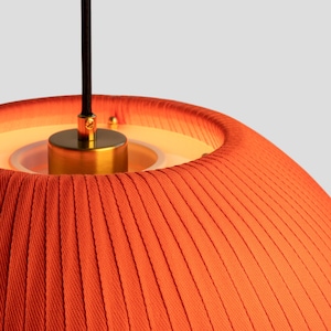 May include: Close-up of a modern orange pendant light. The lampshade has a pleated texture and a warm, inviting glow. The fixture includes a brass-colored metal base and a black cord for hanging. The design is minimalist and stylish.