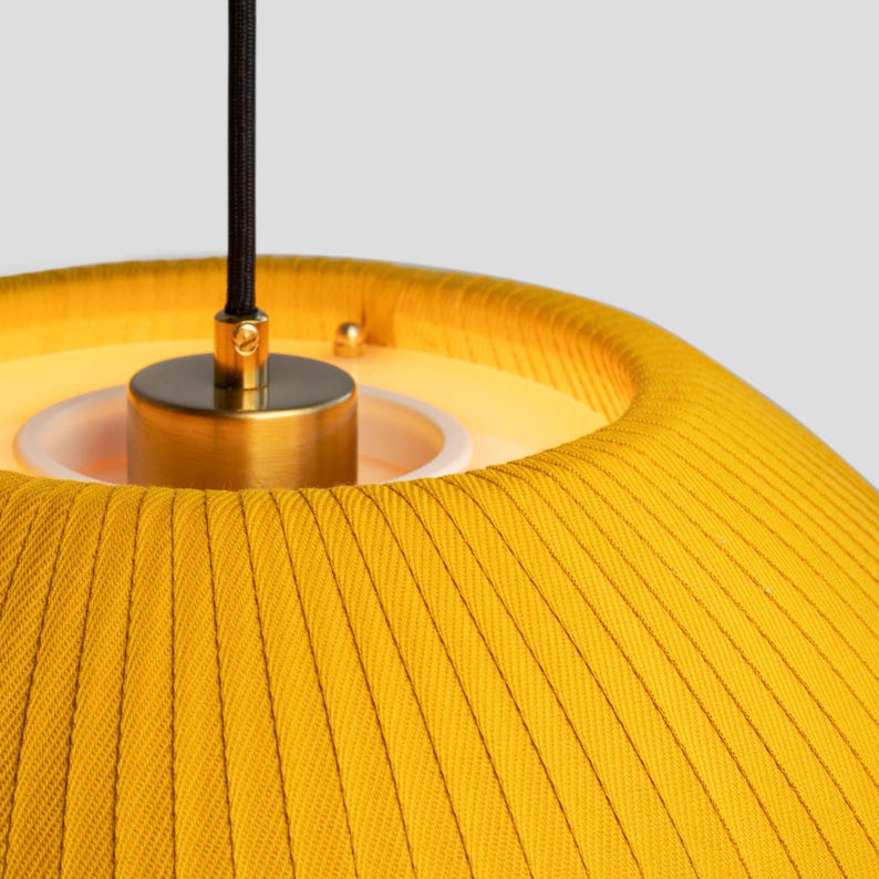 May include: Close-up of a yellow pendant lamp with a pleated fabric shade. The lamp has a brass-colored metal fixture and a black cord. The shade is a vibrant yellow color, and the lamp is lit from within.