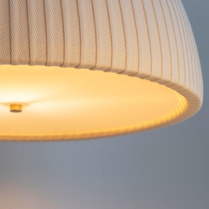 May include: Close-up of a beige fabric lampshade with vertical ribbed detailing. The lampshade has a rounded, inverted bowl shape. The interior of the lampshade is illuminated with a warm, yellow light, and a small, gold-colored fixture is visible.
