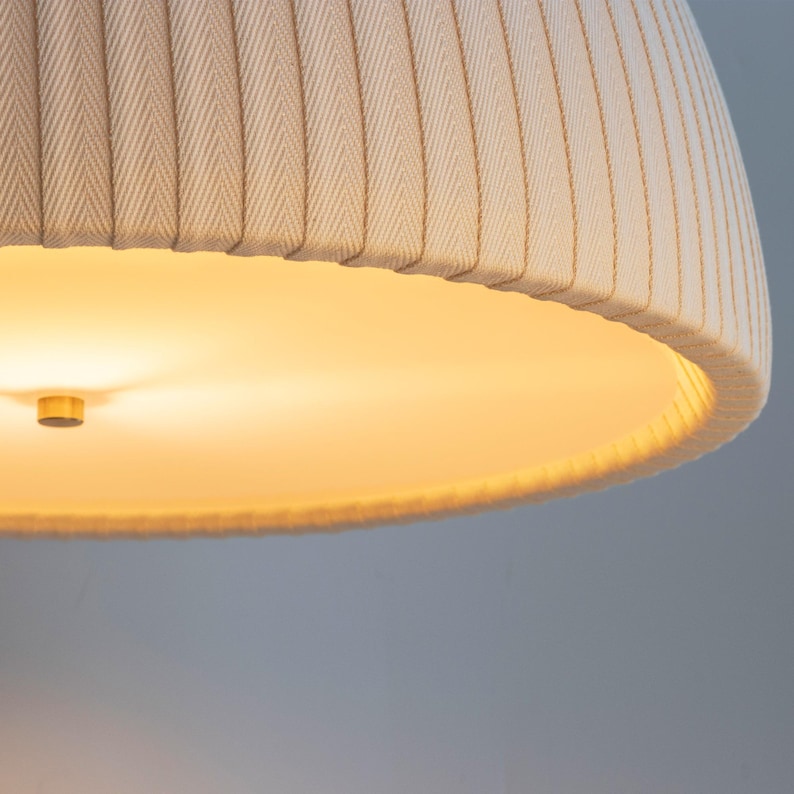May include: Close-up of a beige fabric lampshade with vertical ribbed detailing. The lampshade has a rounded, inverted bowl shape. The interior of the lampshade is illuminated with a warm, yellow light, and a small, gold-colored fixture is visible.