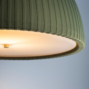 May include: Close-up of a green fabric lampshade with a ribbed texture. The lampshade has a rounded, bell-like shape and a white interior. The light is on, illuminating the interior of the lampshade. The background is a soft blue.