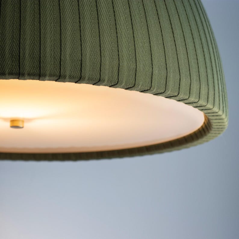 May include: Close-up of a green fabric lampshade with a ribbed texture. The lampshade has a rounded, bell-like shape and a white interior. The light is on, illuminating the interior of the lampshade. The background is a soft blue.