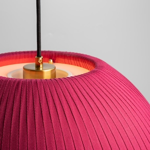 May include: Close-up of a modern pendant light fixture. The lampshade is a deep magenta color with vertical pleats. The fixture has a brass-colored metal base and a black cord. The light source is visible inside the shade.