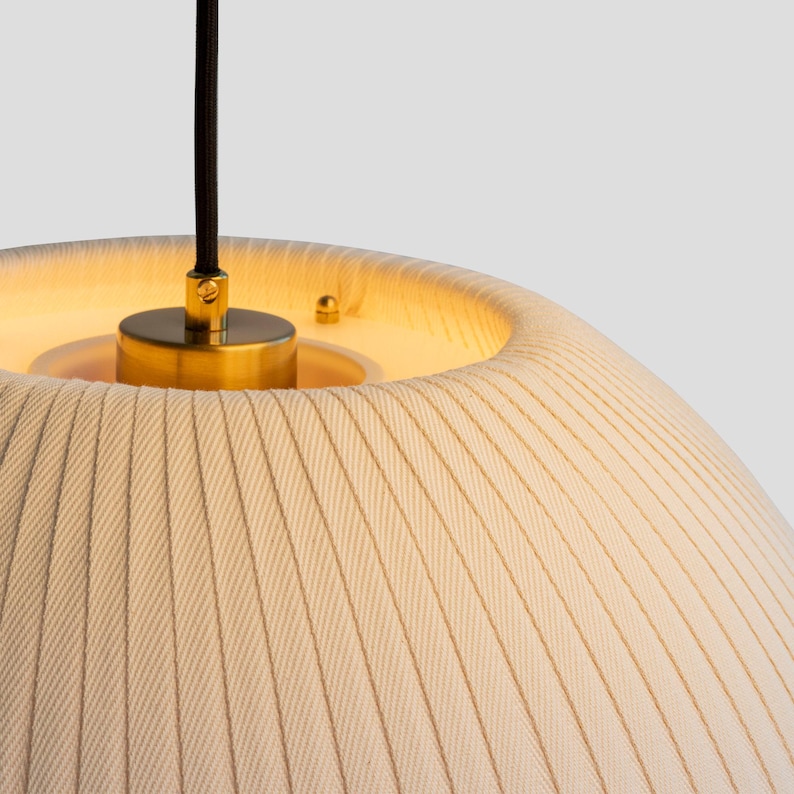 May include: Cream-colored pendant light with a textured, pleated fabric shade. The light fixture has a brass-colored metal base and a black cord. The interior of the shade glows with a warm light.