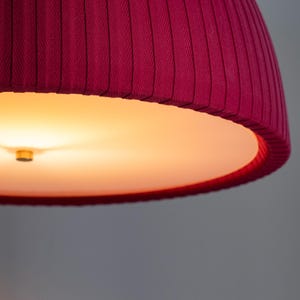 May include: Close-up of a deep magenta lampshade with vertical pleats. The interior is a warm, glowing orange. The lampshade's design features a textured fabric, creating a visually appealing lighting fixture. The overall aesthetic is modern and elegant.