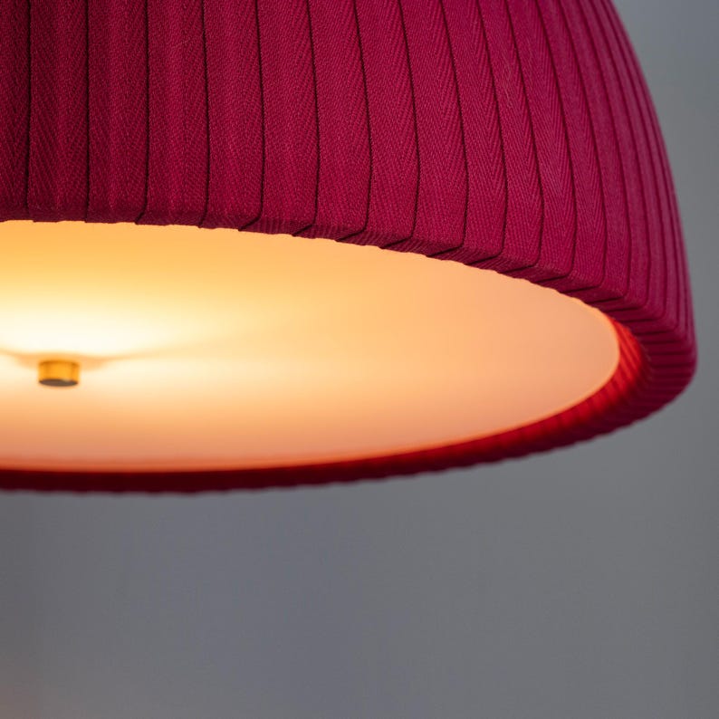 May include: Close-up of a deep magenta lampshade with vertical pleats. The interior is a warm, glowing orange. The lampshade's design features a textured fabric, creating a visually appealing lighting fixture. The overall aesthetic is modern and elegant.