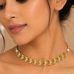 Könnte beinhalten: Ein goldfarbenes Choker-Halskette mit halbmondförmigen Anhängern, die jeweils mit kleinen, weißen Edelsteinen besetzt sind. Die Halskette wird von einer Person mit dunklem Haar und einem Oberteil in neutralen Farben getragen.