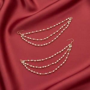 May include: Two gold-coloured earrings, each featuring three strands of small, white, pearl-like beads. The earrings have a curved design and are set against a red fabric backdrop. The earrings are designed to be worn with the hook in the ear and the strands cascading down.