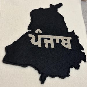 May include: A black felt banner featuring the outline of the Punjab region with the word "ਪੰਜਾਬ" in white lettering. The banner is mounted on a white fabric background.