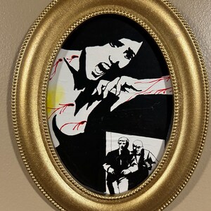 May include: An oval, gold-colored framed artwork. The artwork features a black and white illustration of a screaming figure with red accents. Below the figure is a black and white image of two men.