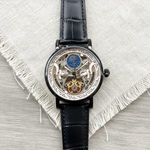 May include: Black skeleton watch with a black leather band. The watch face has a detailed design with gold, red, and blue accents. The watch is displayed on a light gray fabric.