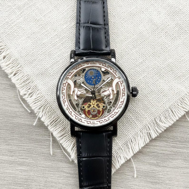 May include: Black skeleton watch with a black leather band. The watch face has a detailed design with gold, red, and blue accents. The watch is displayed on a light gray fabric.