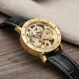 Może przedstawiać: Złoty zegarek szkieletowy z czarnym skórzanym paskiem. Tarcza zegarka odsłania skomplikowane mechanizmy wewnętrzne. Pasek ma białe przeszycia. Zegarek leży na drewnianej powierzchni.