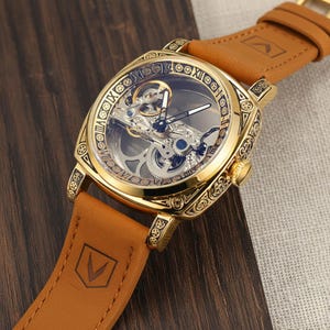 Könnte beinhalten: Goldfarbene, quadratische Armbanduhr mit transparentem Zifferblatt, das komplizierte mechanische Komponenten zeigt. Die Uhr hat römische Ziffern und ein braunes Lederarmband. Auf dem Armband ist ein Logo geprägt.