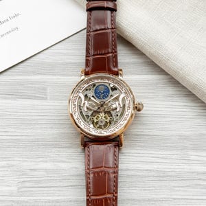 Könnte beinhalten: Eine goldfarbene Skelettuhr mit einem braunen Lederarmband. Das Zifferblatt der Uhr zeigt komplizierte Zahnräder und ein blau-goldenes Himmelsdesign. Das Armband ist etwa 2 cm breit. Die Uhr liegt auf einer hellen Holzmaserung.