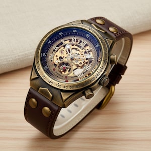 Puede incluir: Un reloj de estilo vintage con una caja de color bronce y una correa de cuero marrón. La esfera del reloj presenta un diseño de esqueleto, mostrando el mecanismo interno. La correa se asegura con remaches y una hebilla dorada.
