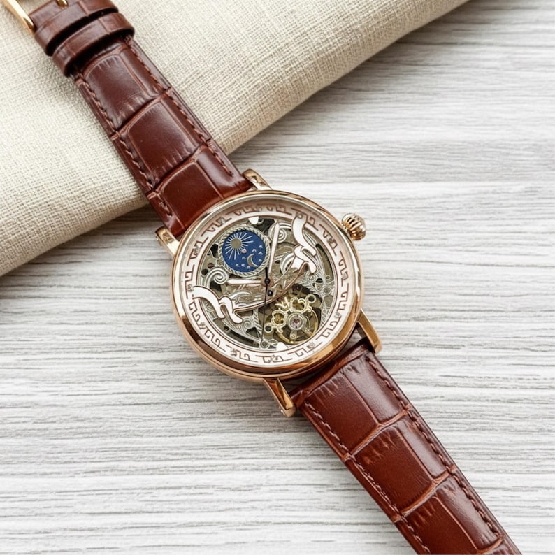 May include: A gold-toned skeleton watch with a brown leather band. The watch face features intricate gears and a blue and gold design. The band has a crocodile pattern.