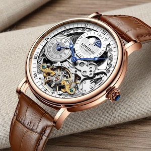 May include: A rose gold automatic wristwatch with a brown leather band. The watch face has a white background with Roman numerals and visible gears. The watch has blue hands and the text "KIMSOUN AUTOMATIC".