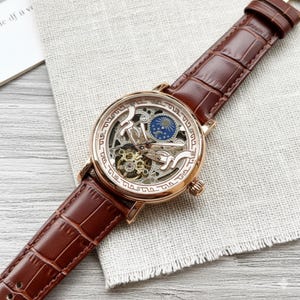 May include: A rose gold skeleton watch with a brown leather band. The watch face features intricate gears and a blue and gold moon phase indicator. The band has a crocodile pattern.