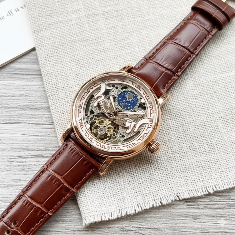 May include: A rose gold skeleton watch with a brown leather band. The watch face features intricate gears and a blue and gold moon phase indicator. The band has a crocodile pattern.